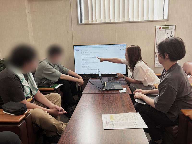 障碍者ITカレッジ愛西の就労支援員・安井よしえが、Google検索結果に表示されたハローワークインターネットサービスの公式サイトを指差し、正しいページへのアクセス方法を訓練生にレクチャーしている様子。