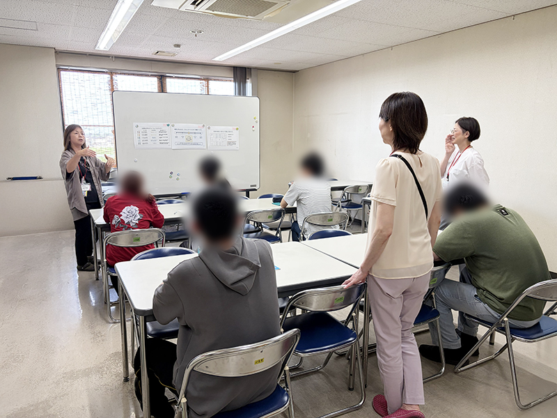 就労支援員による熱心な講義を、フロアの後方から見守るサービス管理責任者の萬谷。訓練生一人ひとりの反応を確かめながら、いつでも些細な相談に応じられる体制を整え、事業所全体で利用者さんの「やってみたい」という気持ちを一番近くで支えている支援風景。