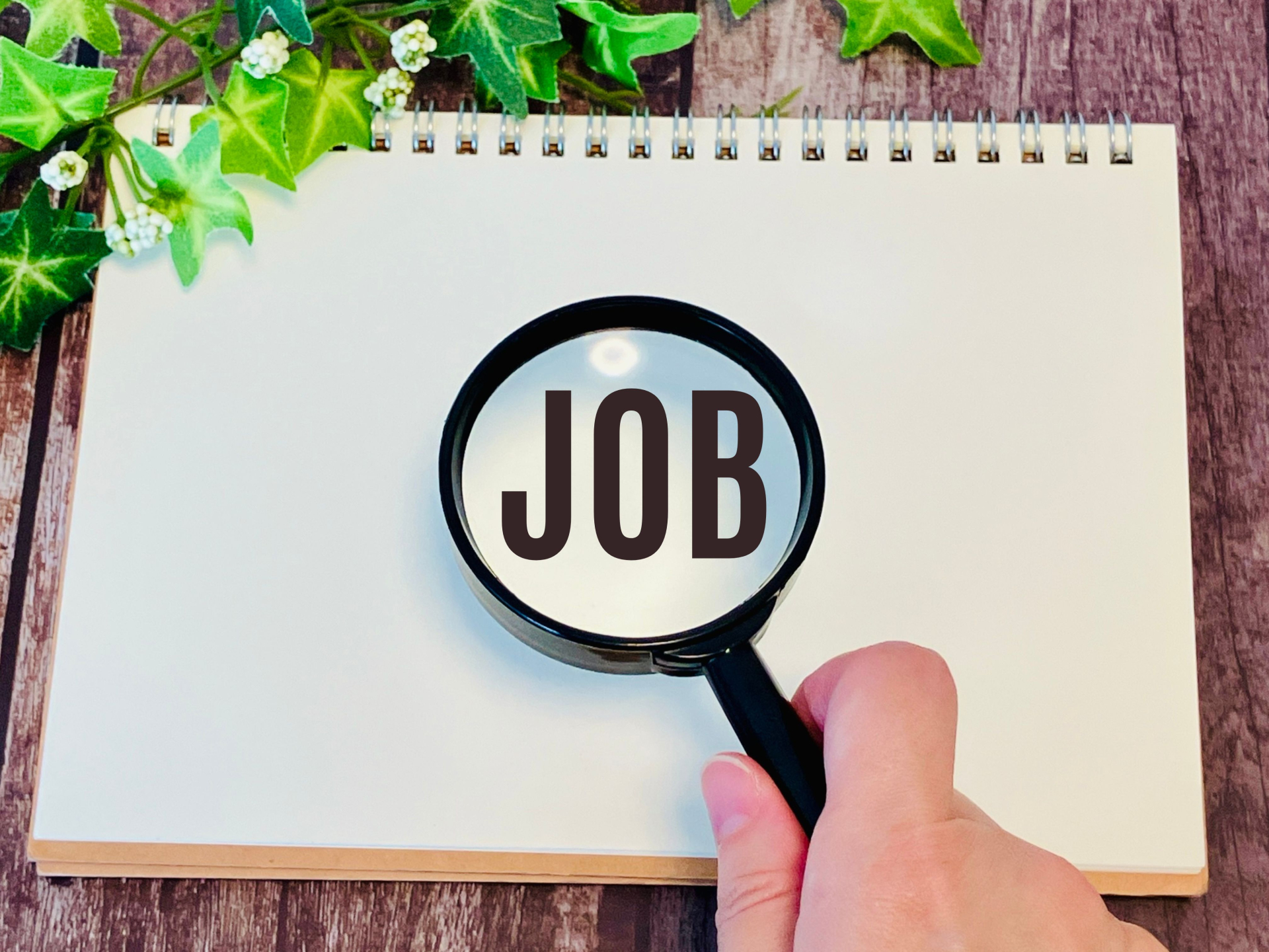 木目調のテーブルの上のノートに書かれた「JOB」の文字を、虫眼鏡（ルーペ）で大きく覗き込んで注意深く拡大している手元の写真。単に書かれた条件を読むだけでなく、自分に合った求人をしっかり見極めて実際の就職活動へつなげていく「求人票の見方」カリキュラムのゴールを表す一枚。