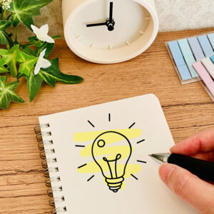 木目調デスクのノートに「ひらめく電球」のイラストが描かれ、ペンを持つ手や付箋、時計が配置されたイメージ画像。グループワークで「人と協力して働くために大切なこと」のアイデアや気づきを出し合うテーマを表現。