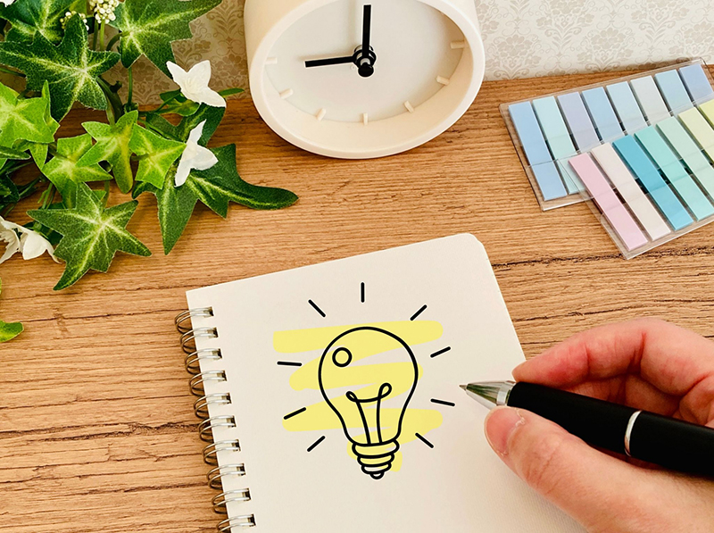 木目調デスクのノートに「ひらめく電球」のイラストが描かれ、ペンを持つ手や付箋、時計が配置されたイメージ画像。グループワークで「人と協力して働くために大切なこと」のアイデアや気づきを出し合うテーマを表現。